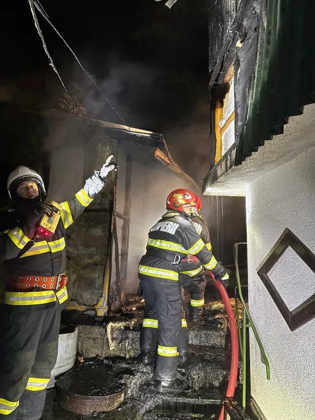 Éjszakai tűzoltási munkálat egy leégett épület mellett: két tűzoltó látható védőfelszerelésben, sisakkal, az egyik piros sisakos tömlővel dolgozik, a másik felfelé mutat egy megrongálódott tetőszerkezet felé, körülöttük füst és megszenesedett gerendák láthatók.