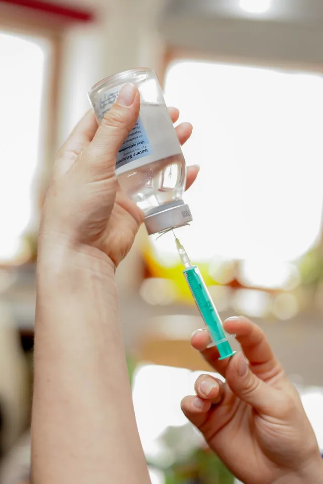 Két kéz látható közelről: az egyik egy fiziológiás sóoldatot (Isotone Natriumchlorid) tartalmazó üvegcsét tart, a másik egy fecskendőt, amellyel a folyadékot szívják ki az üvegcséből.