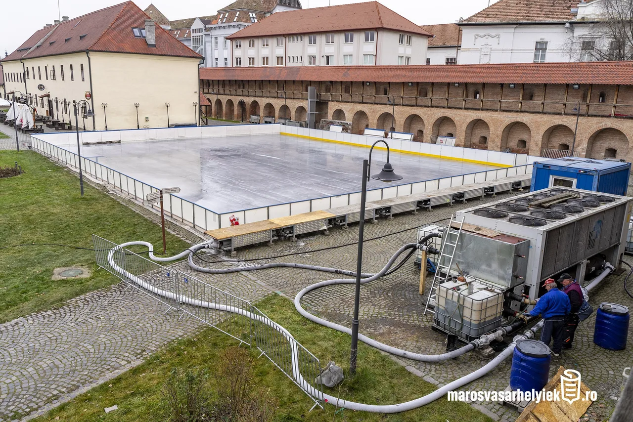 Madártávlati kép a marosvásárhelyi vár udvarán felállított műjégpályáról, körülötte a vár boltíves fala és régi épületek. A jégpálya mellett hűtőberendezés, tömlők és tartályok láthatók, két munkás dolgozik a gépeknél.