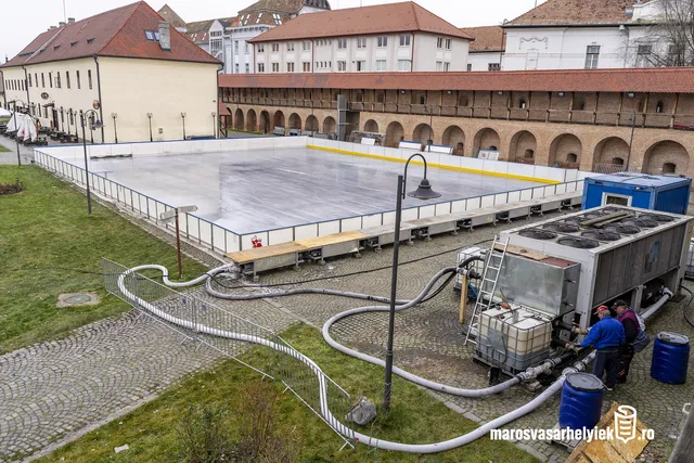 Madártávlati kép a marosvásárhelyi vár udvarán felállított műjégpályáról, körülötte a vár boltíves fala és régi épületek. A jégpálya mellett hűtőberendezés, tömlők és tartályok láthatók, két munkás dolgozik a gépeknél.