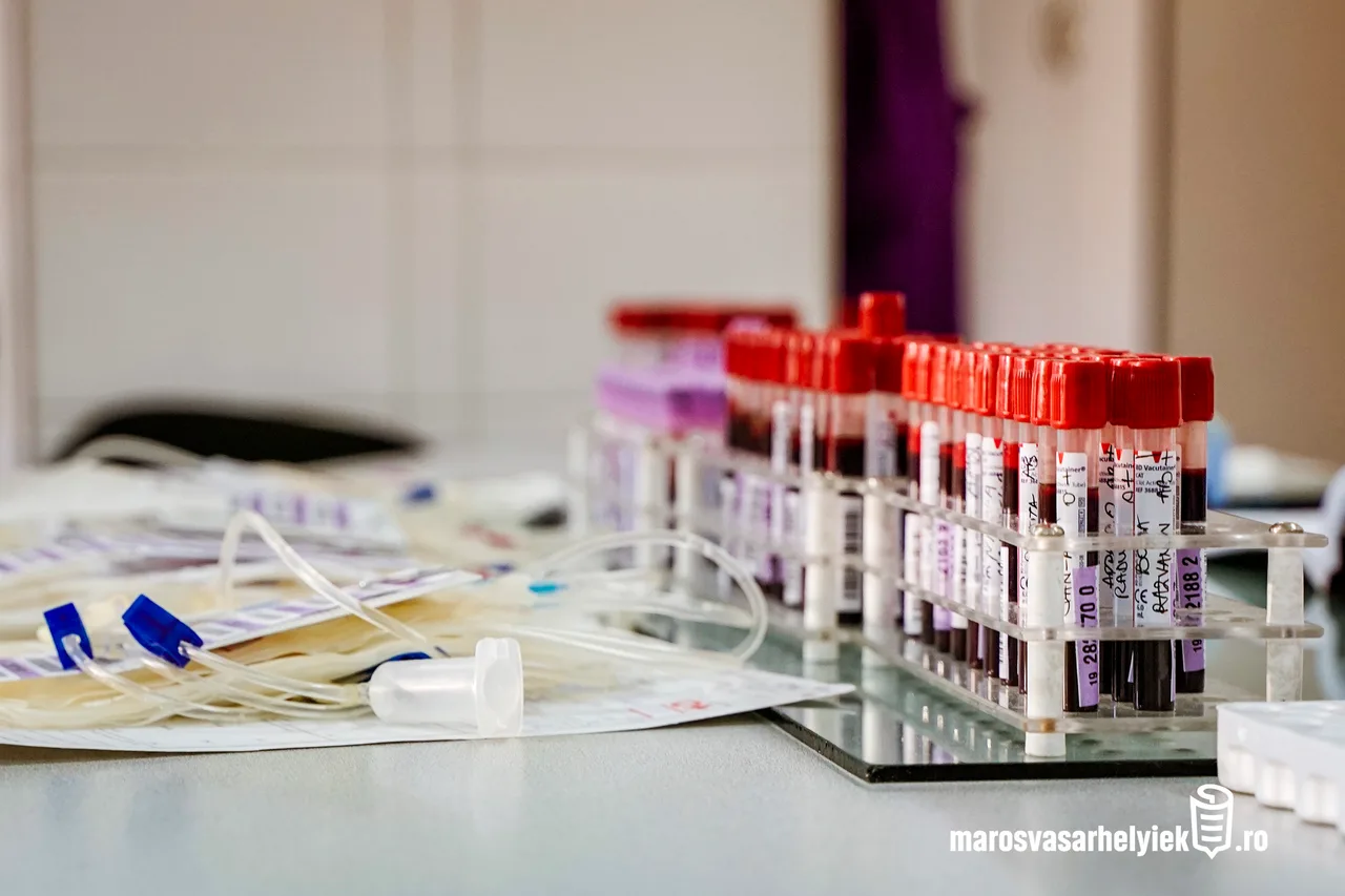 Egy laboratóriumi asztalon vérmintás csövek sorakoznak tartóban, piros kupakokkal, feliratozott címkékkel; előtérben véradáshoz használt zacskók és csövek láthatók.