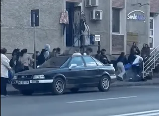 Utcaképen csoportosuló fiatalok egy épület előtti járdán és lépcsőn, egy sötét színű Audi személygépkocsi parkol az úton, a háttérben egy üzlet felirata látszik.