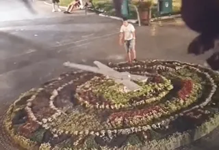Éjszakai utcakép egy kör alakú virágóráról, amelynek mutatóit egy férfi éppen forgatja, körülötte néhány ülő alak látható a háttérben.