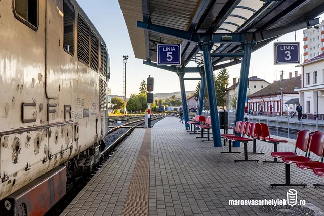 Vasútállomás fedett peronja, a bal oldalon egy régi, kopott CFR mozdony áll a síneken, jobb oldalon piros ülőpadok sorakoznak a fedett várakozóhely alatt. A háttérben a vágányok, jelzőlámpa, valamint a „Linia 5” és „Linia 3” feliratú táblák láthatók, távolabb az állomásépület és a városkép látszik.