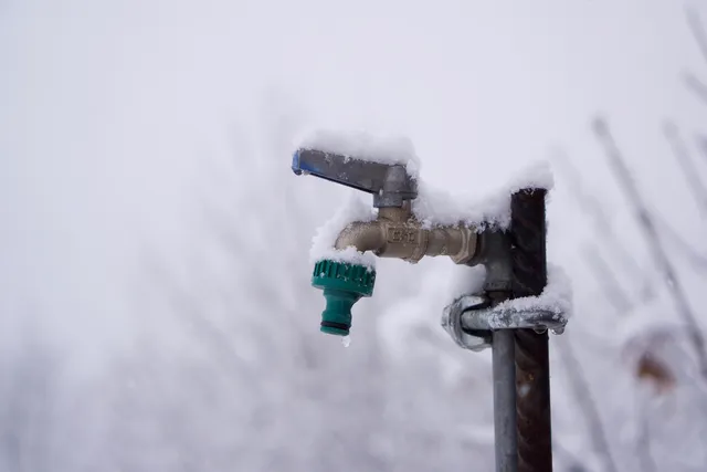 Fémcsapra rögzített kültéri vízcsap zöld locsolócsatlakozóval, amelyet hó és jégcsapok borítanak, a háttérben havas ágak láthatók.