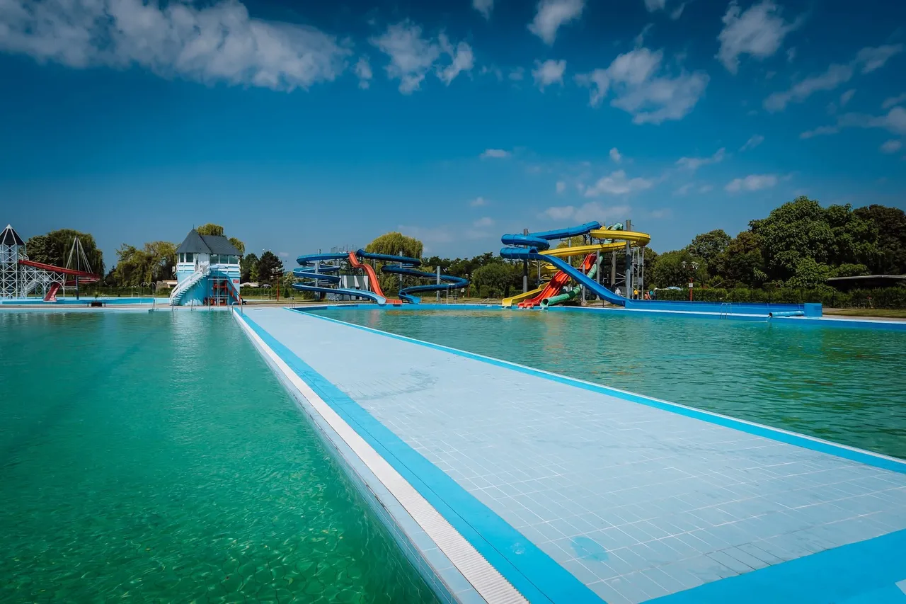Nagy szabadtéri úszómedence, mellette csúszdapark színes csúszdákkal, háttérben fák és kék, felhős égbolt. Az uszoda üres, nincs benne fürdőző.
