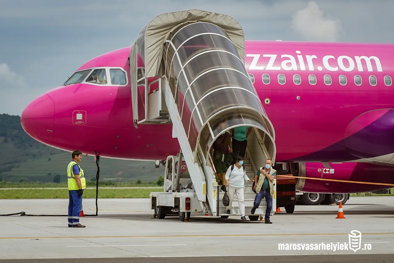 Wizz Air rózsaszín repülőgép áll a repülőtéren, gurulólépcsőn utasok szállnak le, arcmaszkot viselő férfi és nő az előtérben, mellette sárga láthatósági mellényt viselő földi kiszolgáló személyzet tagja.