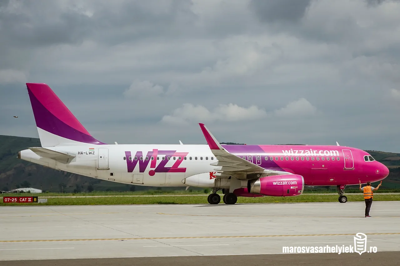 Wizz Air rózsaszín-fehér repülőgép (HA-LWZ) áll a repülőtéri gurulóúton, mellette egy jelzőmellényes földi személyzet tagja irányítja a gépet, a háttérben felhős égbolt és dombok láthatók.