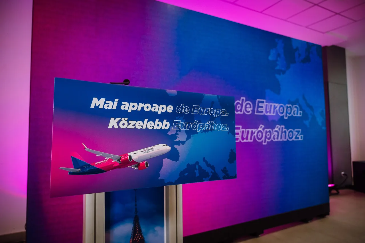 Egy nagy kivetítőn és egy előtte álló táblán a Wizz Air reklámja látható, rajta repülőgép és Európa térképe, felirat: „Mai aproape de Europa. Közelebb Európához.” A háttérben rózsaszín és kék fények világítják meg a termet.
