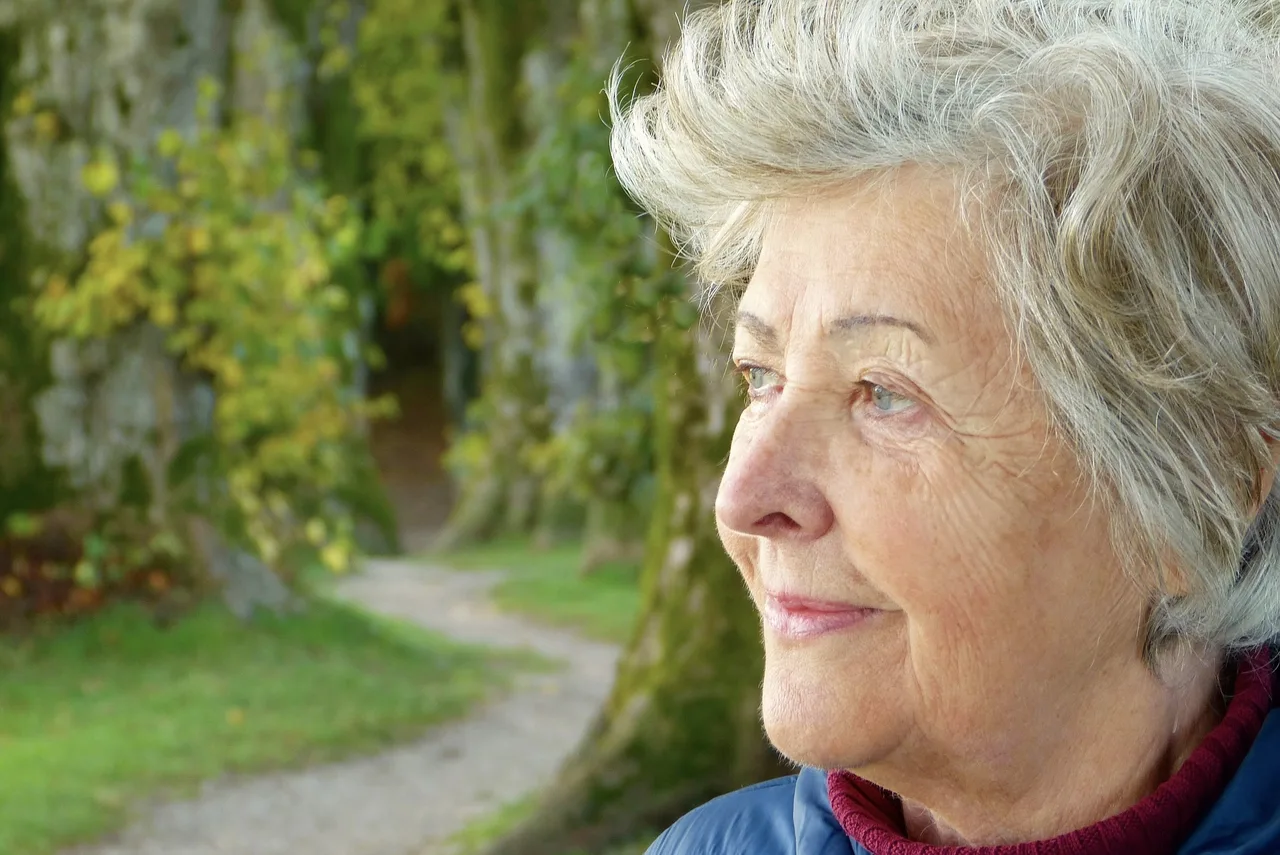 Idős, ősz hajú nő közelről fotózva, oldalról néz a távolba, mögötte fákkal szegélyezett park és kavicsos ösvény látszik.