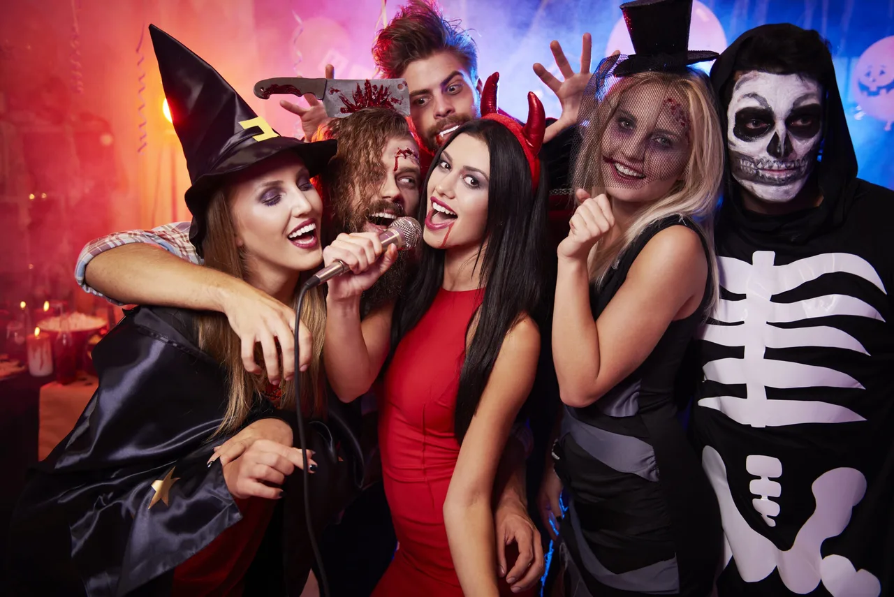 Hat fiatal felnőtt halloweeni jelmezben pózol egy mikrofonnal a kezükben, boszorkány, ördög, koponyás-vázlatos, vérző sebhelyes és kaszás jelmezekben, színes fényekkel megvilágított háttér előtt.